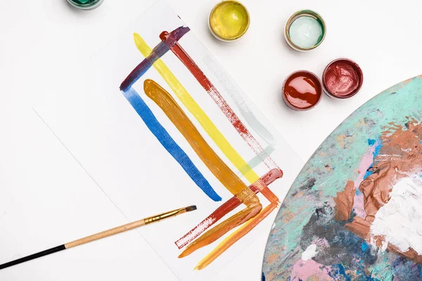 Vue du dessus de la palette avec peintures Gouache et pinceau près du papier avec coups de pinceau sur fond blanc — Photo de stock