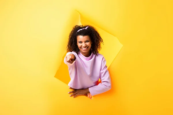 Cheerful african american woman pointing with finger through ripped paper on yellow — Stock Photo