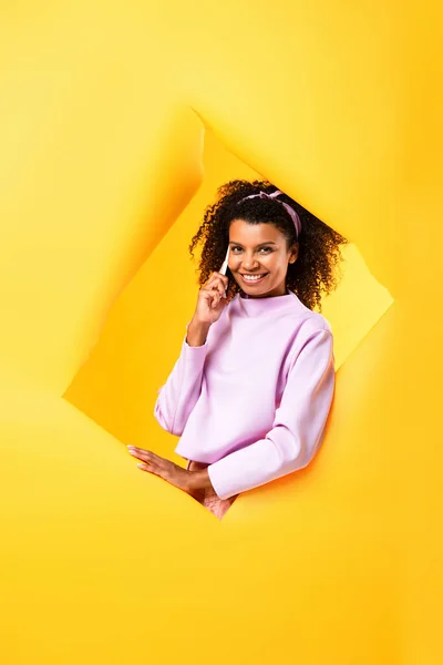 Cheerful african american woman talking on smartphone and looking at camera through ripped paper on yellow — Stock Photo