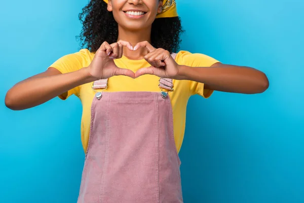 Vista recortada de la alegre mujer afroamericana mostrando el signo del corazón con las manos aisladas en azul - foto de stock