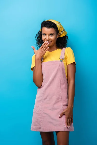 Tímida africana americana mujer cubriendo la boca con la mano aislada en azul - foto de stock