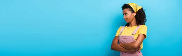 Mujer afroamericana escéptica de pie con los brazos cruzados y mirando hacia otro lado en azul, bandera - foto de stock