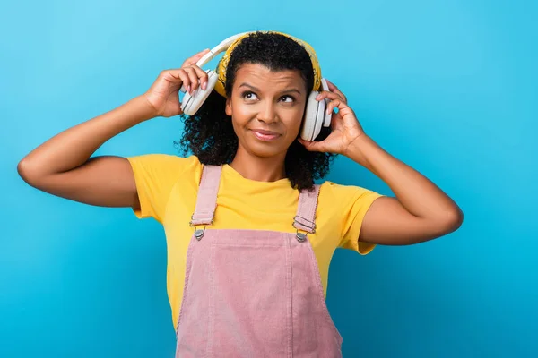 Femme afro-américaine confus dans les écouteurs sans fil écouter de la musique sur bleu — Photo de stock