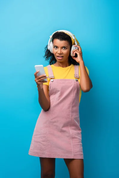 Femme afro-américaine choquée dans les écouteurs écouter de la musique et en utilisant un smartphone sur bleu — Photo de stock