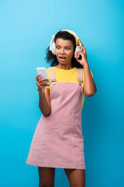 Shocked african american woman in wireless headphones listening music and using smartphone on blue — Stock Photo