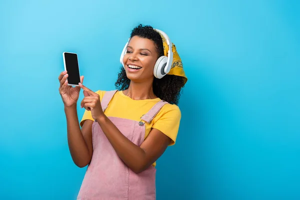 Mujer afroamericana feliz en auriculares escuchando música y señalando con el dedo al teléfono inteligente con pantalla en blanco en azul - foto de stock