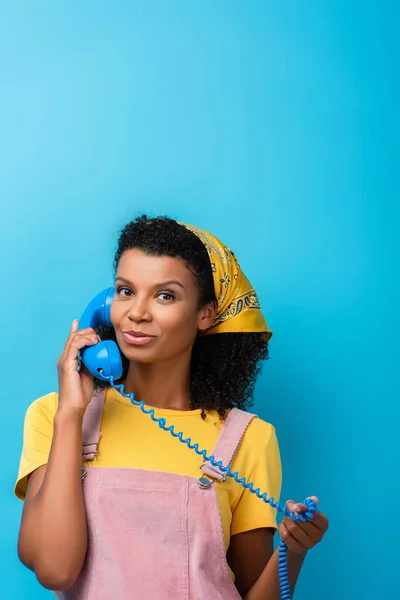 Curly african american woman talking on retro telephone and looking at camera on blue — Stock Photo