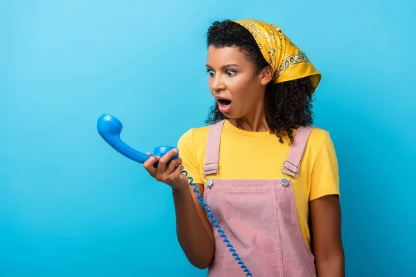 Impactado mujer afroamericana mirando retro teléfono en azul - foto de stock