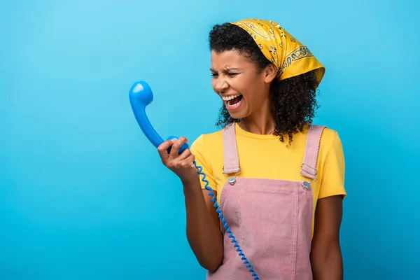 African american woman looking at retro telephone and screaming on blue — Stock Photo