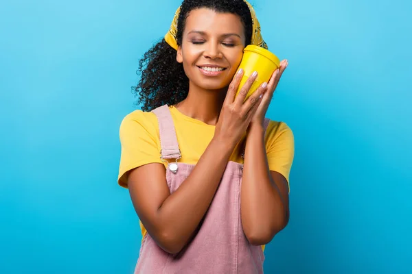 Mujer afroamericana feliz con los ojos cerrados sosteniendo taza reutilizable en azul - foto de stock