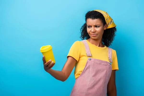 Mujer afroamericana escéptica sosteniendo taza reutilizable en azul - foto de stock