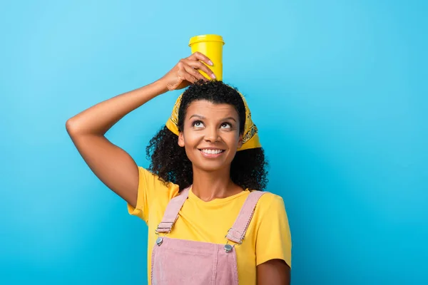 Mujer afroamericana feliz sosteniendo taza reutilizable por encima de la cabeza en azul - foto de stock