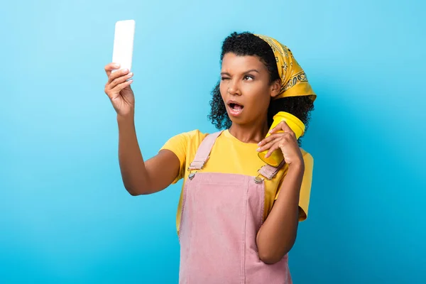 Mujer afroamericana guiñando el ojo, sosteniendo taza reutilizable y tomando selfie en azul - foto de stock