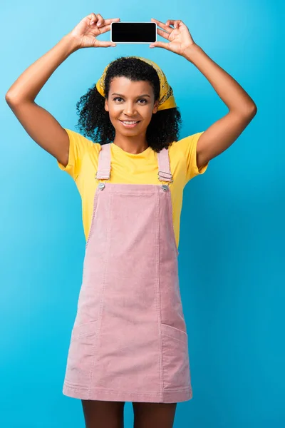 Heureuse femme afro-américaine tenant smartphone avec écran vide au-dessus de la tête sur bleu — Photo de stock