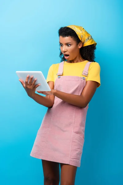 Femme afro-américaine choquée en foulard en utilisant une tablette numérique sur bleu — Photo de stock