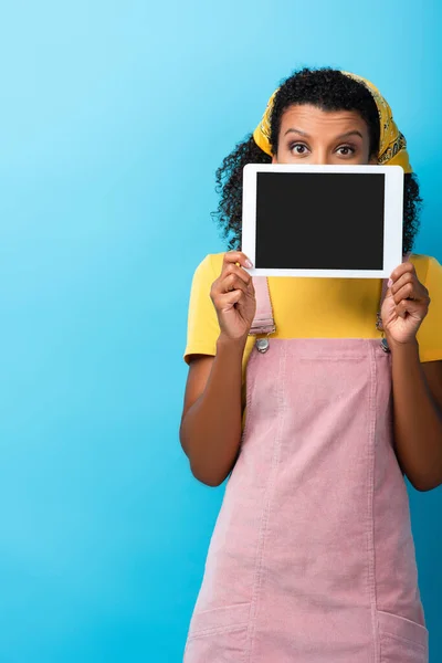 Mujer afroamericana rizada cubriendo la cara mientras sostiene la tableta digital con pantalla en blanco en azul - foto de stock