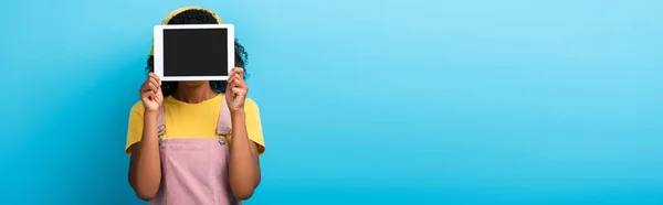 Femme afro-américaine bouclée couvrant le visage tout en tenant tablette numérique avec écran blanc sur bleu, bannière — Photo de stock