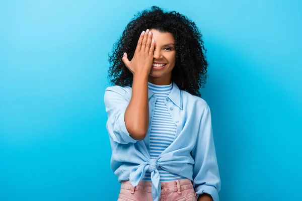 Кучерява афроамериканська жінка прикриває очі і посміхається на синьому — стокове фото