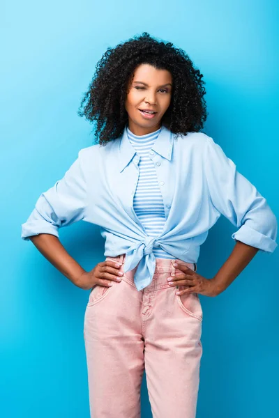 Curly african american woman standing with hand on hips and winking eye on blue — Stock Photo