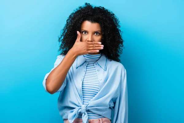 Curly african american woman covering mouth on blue — Stock Photo