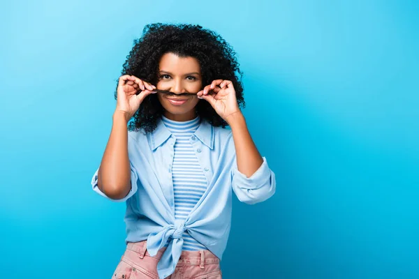 African american woman holding hair near mouth on blue — Stock Photo