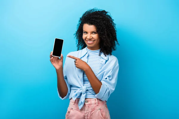 Mujer afroamericana feliz apuntando con el dedo al teléfono inteligente con pantalla en blanco en azul - foto de stock