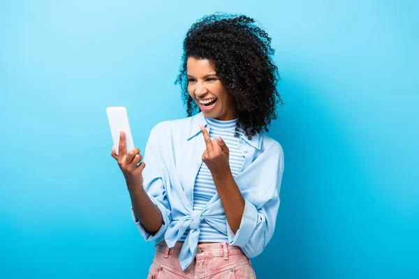 Mulher americana africana feliz mostrando dedo médio para smartphone em azul — Fotografia de Stock