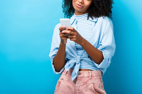 Vista recortada de la mujer afroamericana mensajes de texto en el teléfono inteligente en azul - foto de stock