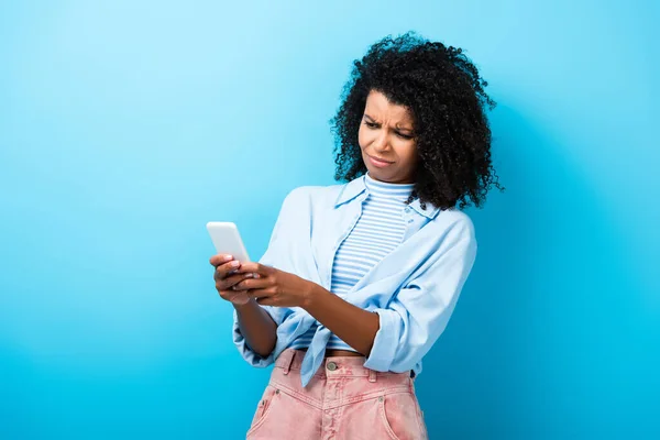 Незадоволена афроамериканська жінка смс на смартфоні на синьому — стокове фото