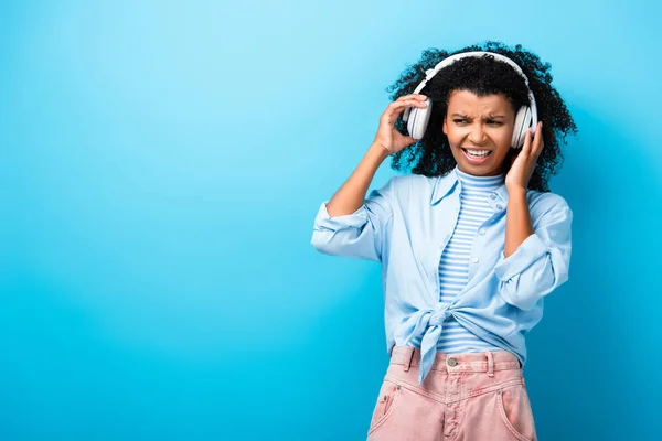 Displeased african american woman listening music in headphones on blue — Stock Photo