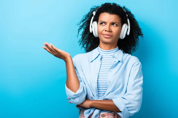 Femme afro-américaine sceptique écouter de la musique dans les écouteurs sur bleu — Photo de stock