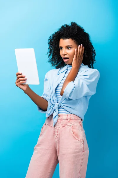 Überraschte Afroamerikanerin blickt auf digitales Tablet auf blau — Stockfoto