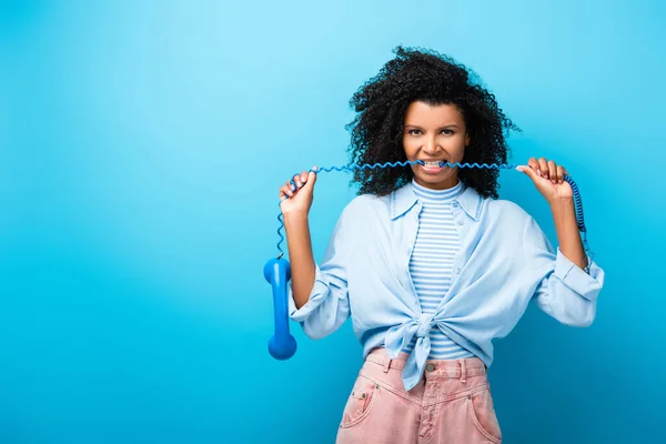 Curly african american woman biting cable on vintage telephone on blue — Stock Photo