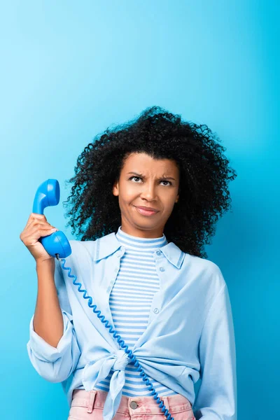 Femme afro-américaine mécontente tenant un téléphone rétro sur bleu — Photo de stock