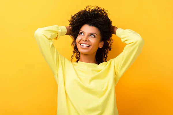 Happy african american woman looking away while fixing curly hair on yellow — Stock Photo