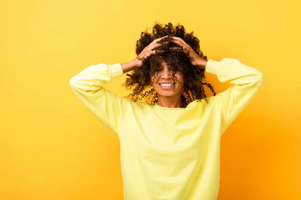 Happy african american woman with closed eyes while fixing curly hair on yellow — Stock Photo