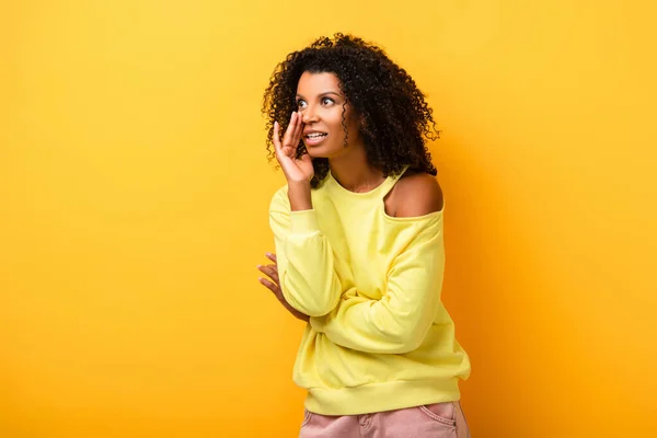 Femme afro-américaine choquée chuchotant sur jaune — Photo de stock