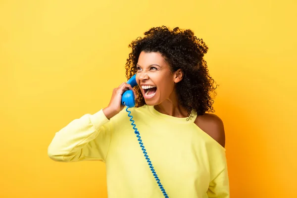 Curly african american woman laughing while talking on vintage telephone on yellow — Stock Photo
