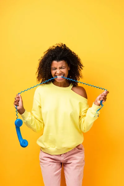 Angry african american woman biting cable of vintage telephone on yellow — Stock Photo