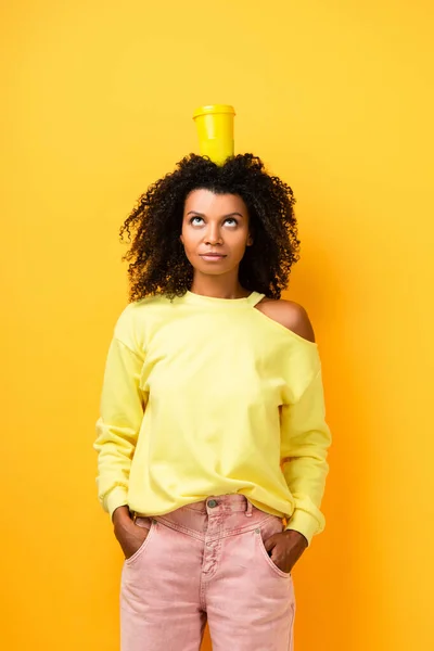 African american woman with reusable cup on head standing with hands in pockets on yellow — Stock Photo