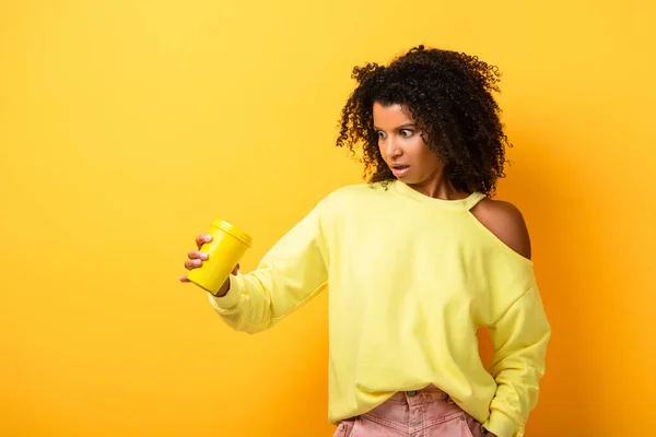 Chocado Africano americano mulher segurando copo reutilizável no amarelo — Fotografia de Stock
