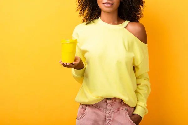 Vista ritagliata della donna afro-americana che tiene in mano la tazza riutilizzabile mentre in piedi con la mano in tasca sul giallo — Foto stock