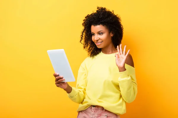 Afro-américaine femme tenant tablette numérique et agitant la main tout en ayant chat vidéo sur jaune — Photo de stock