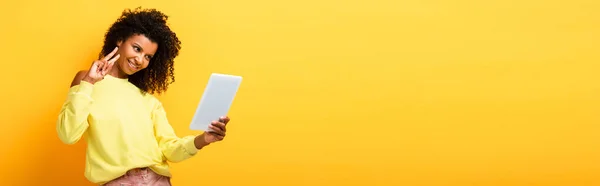 Cheerful african american woman holding digital tablet and showing peace sign on yellow, banner — Stock Photo