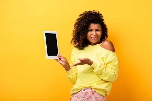 Mujer afroamericana escéptica apuntando con la mano a la tableta digital con pantalla en blanco en amarillo - foto de stock