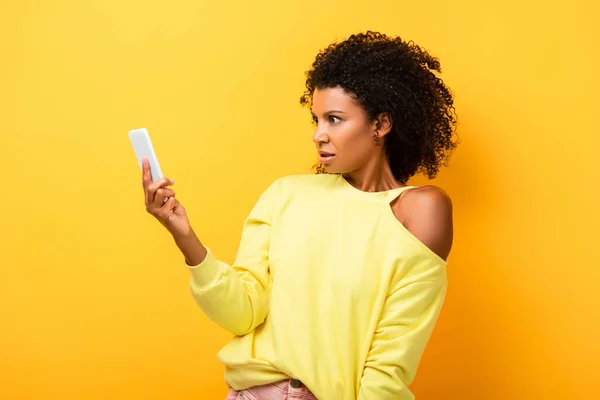 Здивована афроамериканська жінка тримає смартфон на жовтому — стокове фото