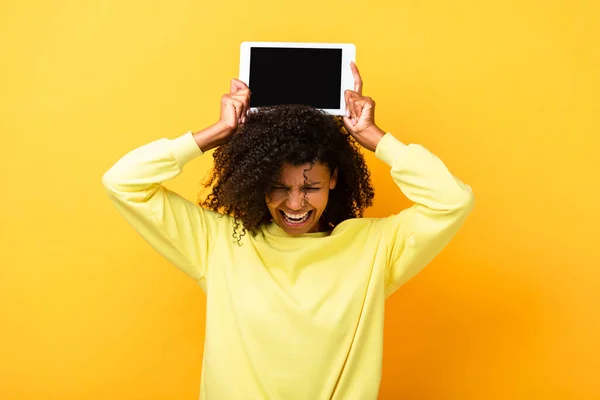 Afro-américaine femme tenant tablette numérique avec écran vierge au-dessus de la tête et criant sur jaune — Photo de stock