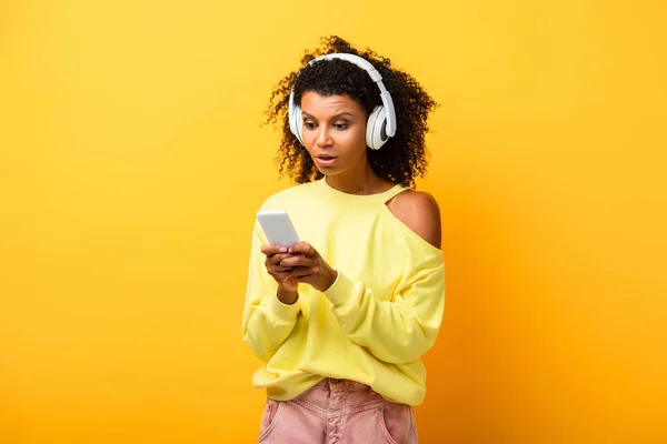 Mulher afro-americana surpreso em fones de ouvido usando smartphone no amarelo — Fotografia de Stock