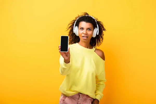 Shocked african american woman in headphones holding smartphone with blank screen on yellow — Stock Photo