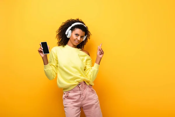 Mujer afroamericana feliz en auriculares que sostienen el teléfono inteligente con la pantalla en blanco mientras que señala con el dedo en amarillo - foto de stock